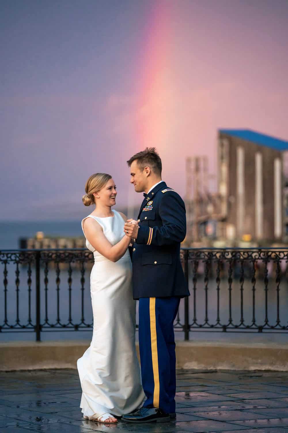 A bride in a white dress and a groom in a military uniform dance on a waterfront esplanade with a colorful sunset sky and rainbow in the background.