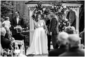 A bride wipes a tear while holding hands with the groom during an outdoor wedding ceremony in front of a decorated gazebo. The wedding party and guests are nearby.