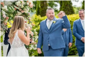 A groom raises his fist in celebration during an outdoor wedding ceremony, next to a bride in a white dress. A floral arrangement and greenery are in the background.