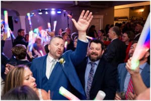 A lively group of people in formal attire are celebrating by waving glow sticks and dancing. One man in a blue suit is prominently raising his hand and singing.