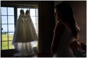 A woman stands indoors adjusting the strap of her white dress, while a wedding dress hangs in the background, illuminated by natural light from a nearby window.