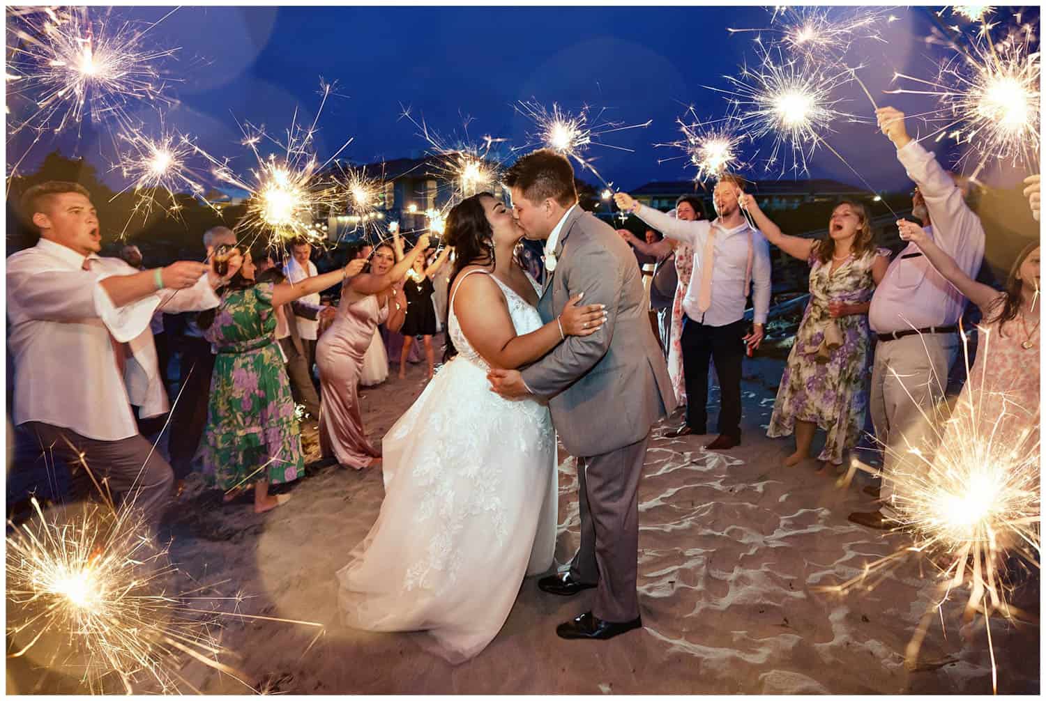 A bride and groom kiss while surrounded by guests holding sparklers at an outdoor nighttime celebration.