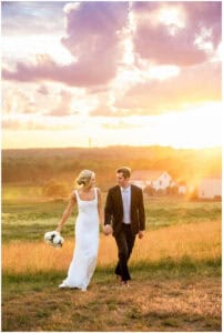 A couple dressed in wedding attire walks hand in hand through a field at sunset.