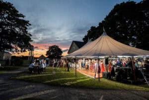 Outdoor evening wedding reception under a large tent with people dancing, socializing, and enjoying the event at sunset.