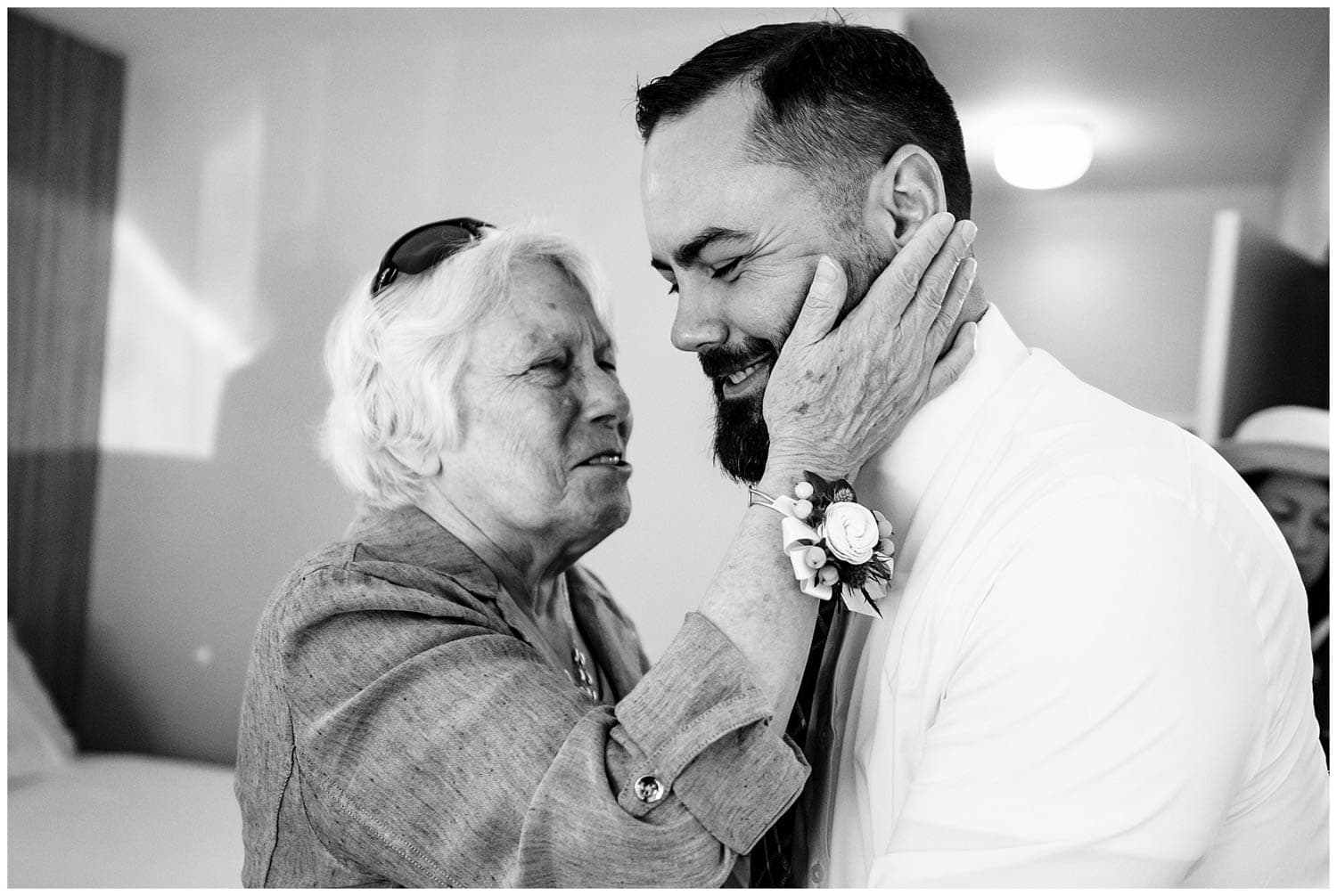 An elderly woman, tenderly touches the bearded face of a groom, who is smiling with his eyes closed. Both appear to be sharing a heartfelt moment indoors.