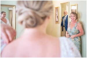 A bride in a white dress stands with her back to the camera while an older man in a suit and a woman in a floral dress smile at her in a room with mirrors.