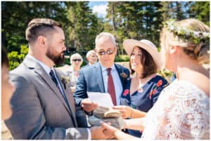 A couple holds hands while a man reads from a paper during an outdoor wedding ceremony; another woman stands beside them, and guests watch in the background.