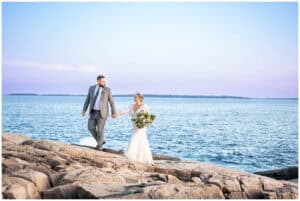 A bride and groom walk on rocky terrain by the sea. The bride holds a bouquet of flowers, and the groom is dressed in a grey suit.
