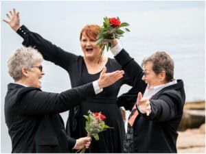 Three women, dressed in black and white attire, celebrate joyfully by the water, holding flowers and extending their arms towards each other.