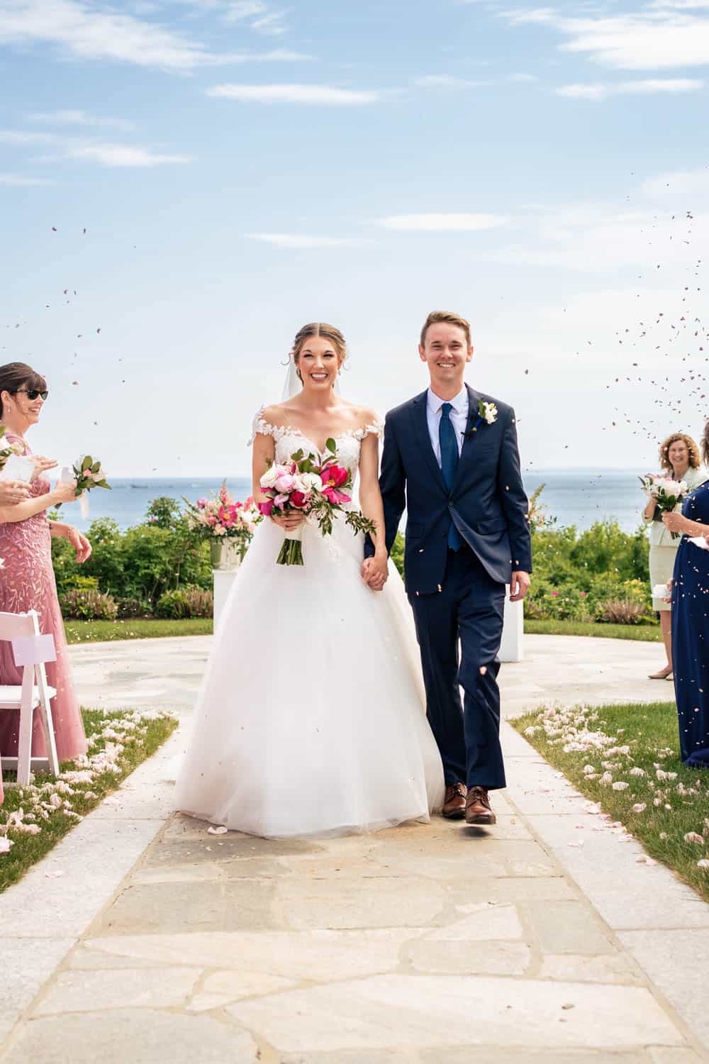 A bride in a white gown and a groom in a navy suit holding hands and walking down an aisle outdoors, with petals being thrown around them and guests on either side.