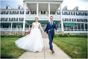 A bride and groom smile as they walk hand in hand on a pathway in front of a large, white, multi-story building, with the bride holding a pink bouquet.