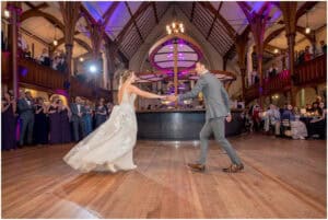 A bride and groom dance in the center of a large, elegantly decorated hall, with guests watching from both the floor and an upper balcony.