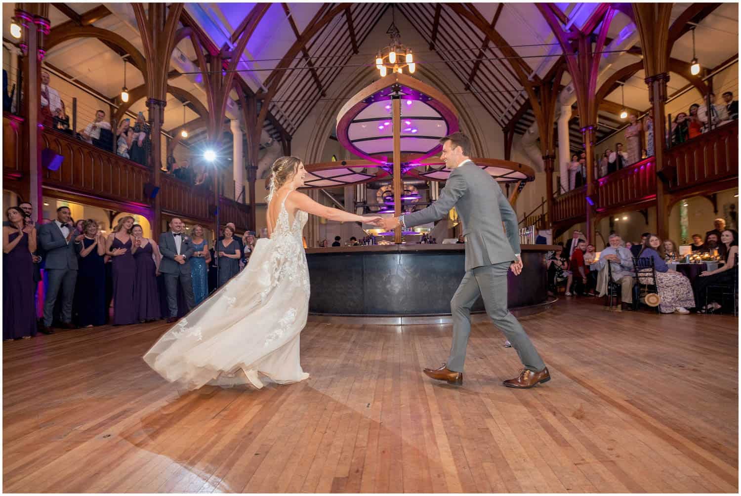 A bride and groom dance in the center of a large, elegantly decorated hall, with guests watching from both the floor and an upper balcony.