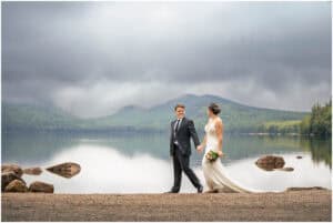 A bride and groom walk hand in hand by a lake with a misty mountain backdrop. The bride holds a bouquet and is wearing a white dress, while the groom is in a suit.