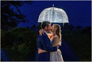 A couple embraces and kisses under a clear umbrella at night. It's raining, and the background is dark with faint outlines of trees and foliage.