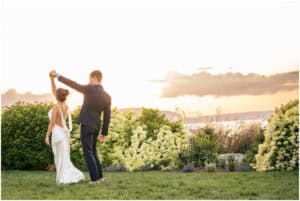 A couple dances on a grassy area near flowering bushes at sunset, the bride in a white dress and the groom in a dark suit.