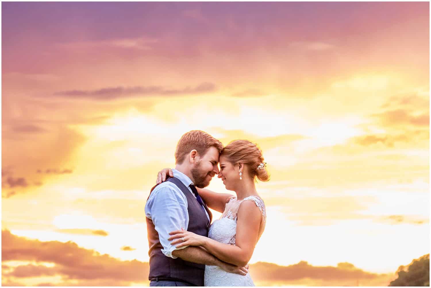 A couple embraces, touching foreheads, standing outside at sunset with a scenic sky in the background.