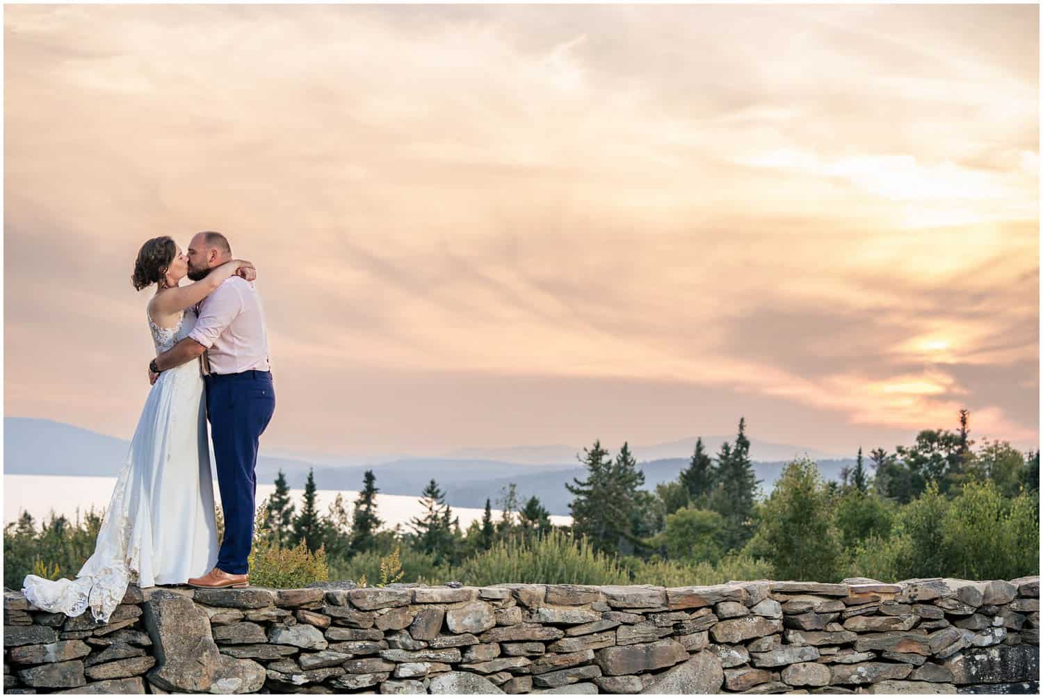 A couple in wedding attire embraces atop a stone wall, with a scenic sunset and trees in the background.