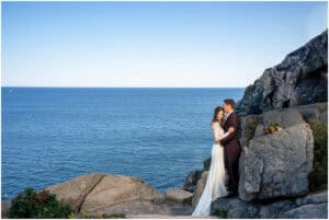 A bride and groom stand on rocky terrain by the sea, embracing each other. The ocean and a clear blue sky form the backdrop.