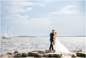 A bride and groom stand on a rocky shore, embracing and kissing, with a sailboat and calm ocean waters in the background under a clear sky.