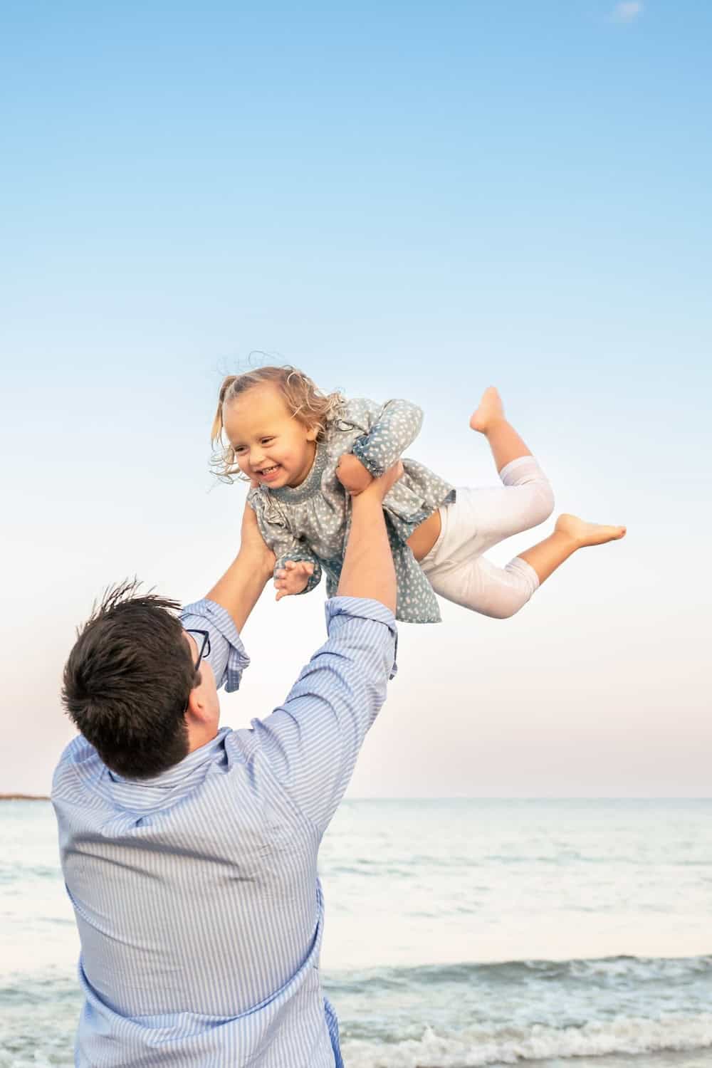 An adult lifts a smiling child into the air on a beach with calm water and a clear sky in the background.