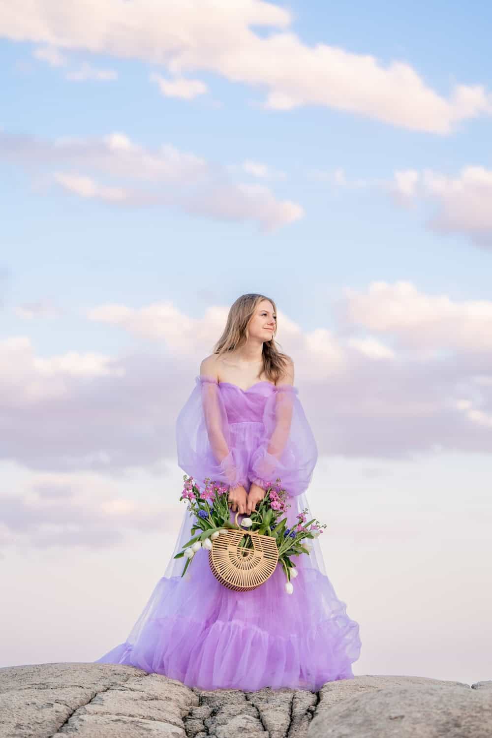 A woman in a lavender dress stands on a rocky surface holding a round wooden basket filled with flowers. The sky in the background is partly cloudy.