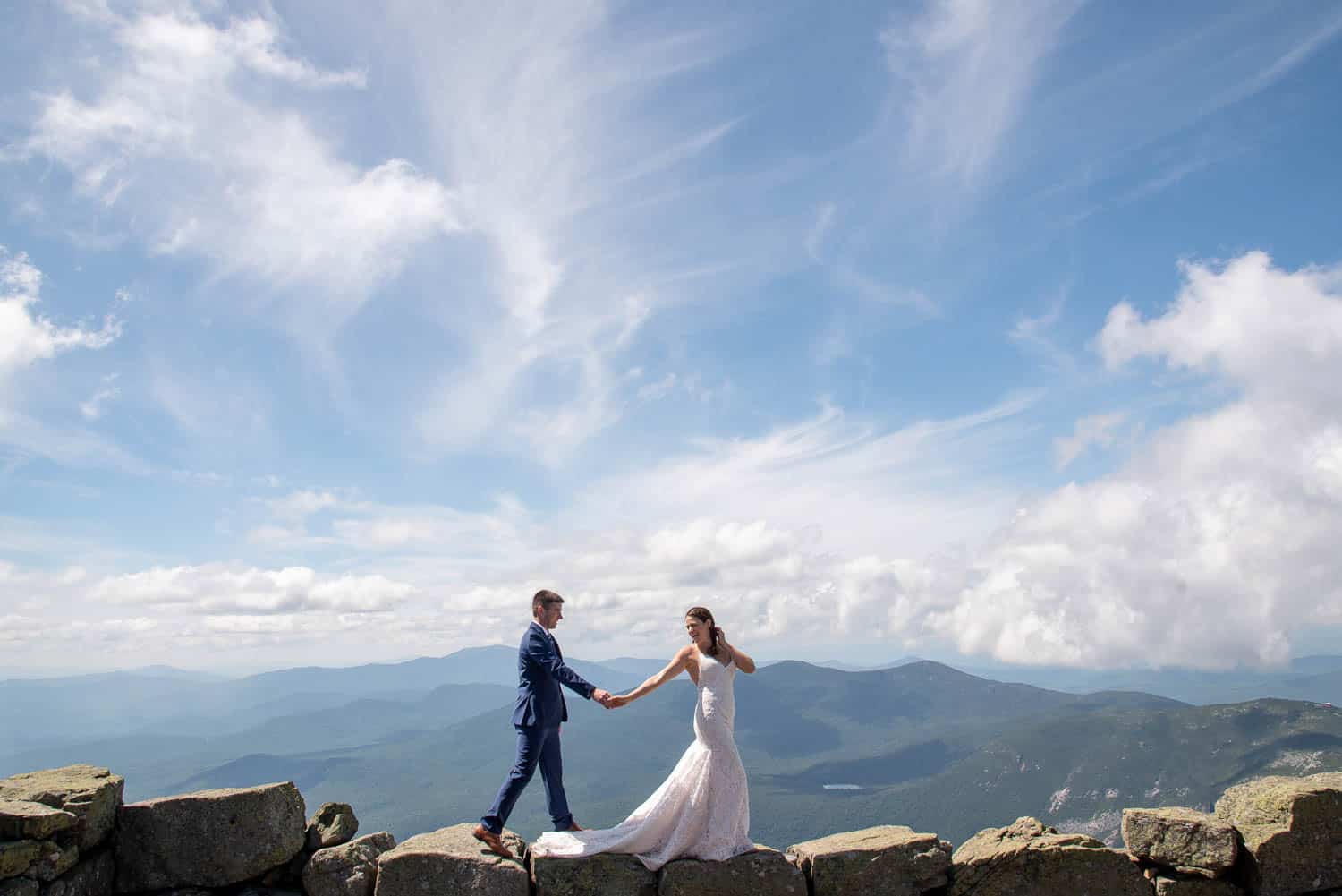 A bride and groom hold hands while standing on a rocky ledge with a vast mountain range and blue sky with scattered clouds in the background.