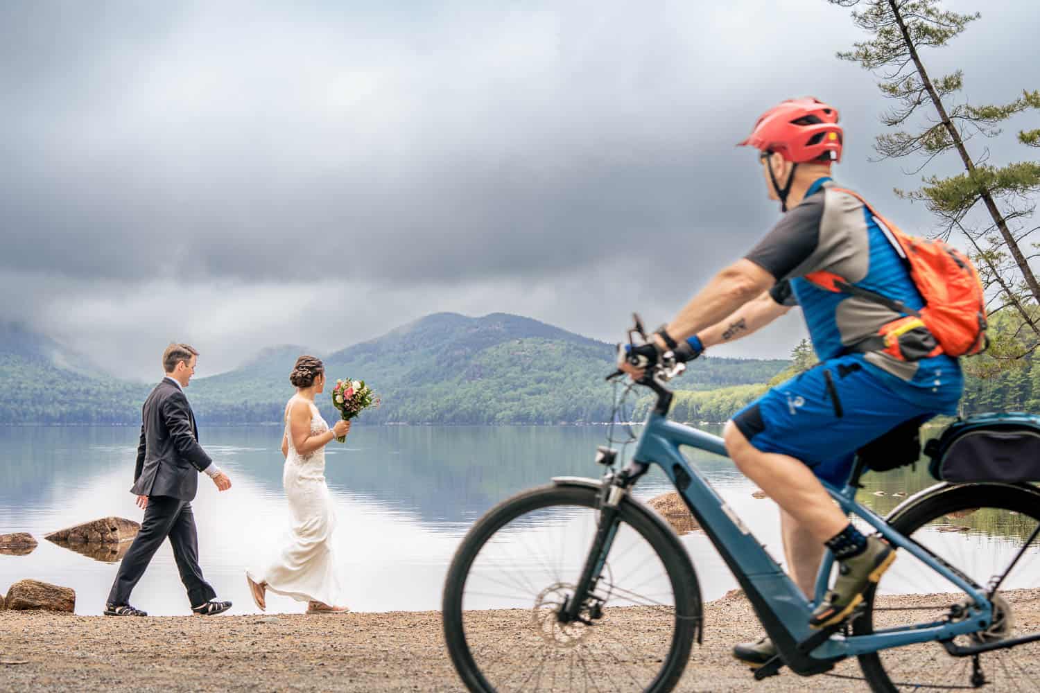 A bride and groom walk hand in hand along a lakeside path, while a cyclist wearing a red helmet rides by. The background features mountains and a cloudy sky.