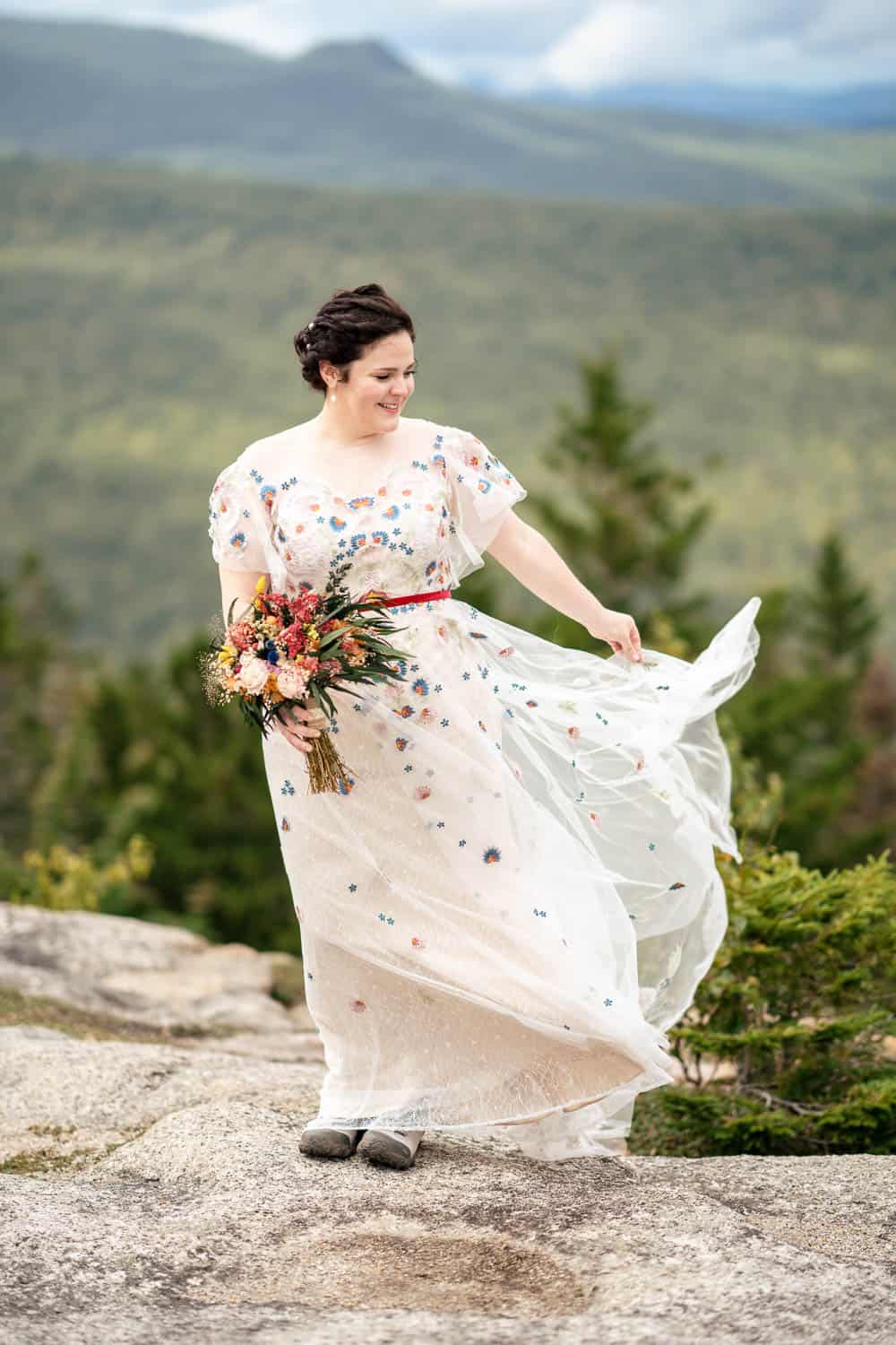 A woman in a white floral dress stands on a rocky outcrop holding a bouquet of flowers with a scenic green mountain landscape in the background.