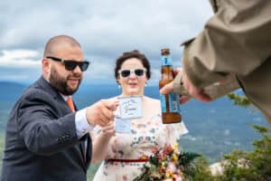 A man and woman dressed formally, wearing sunglasses, are exchanging drinks with another person at an outdoor location. The man holds a cup marked Groom.