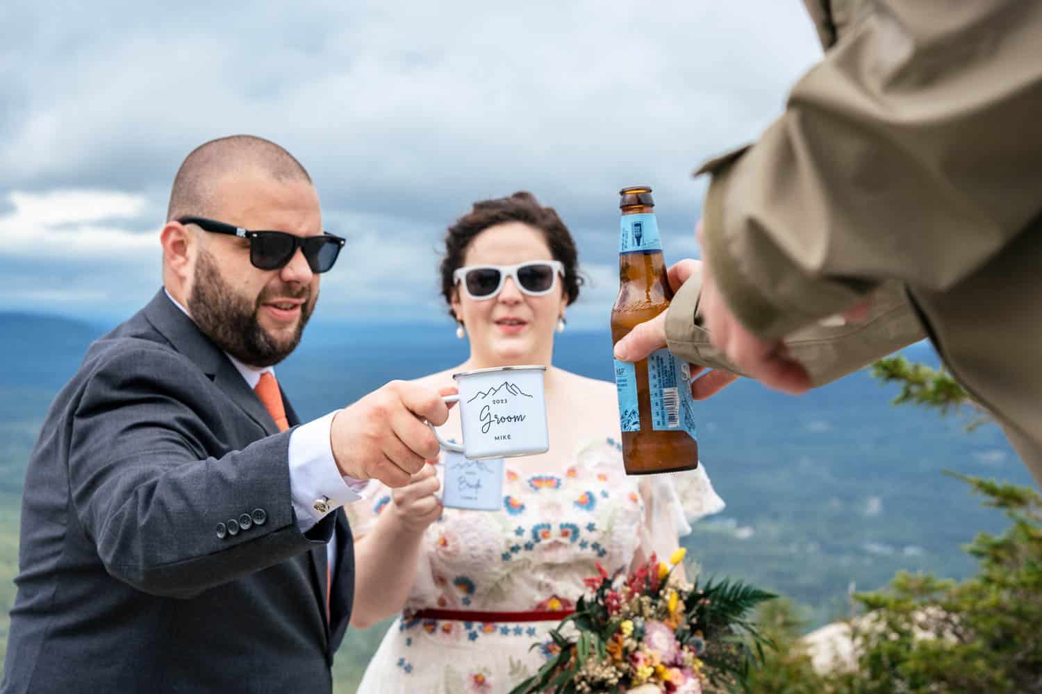 A man and woman dressed formally, wearing sunglasses, are exchanging drinks with another person at an outdoor location. The man holds a cup marked Groom.