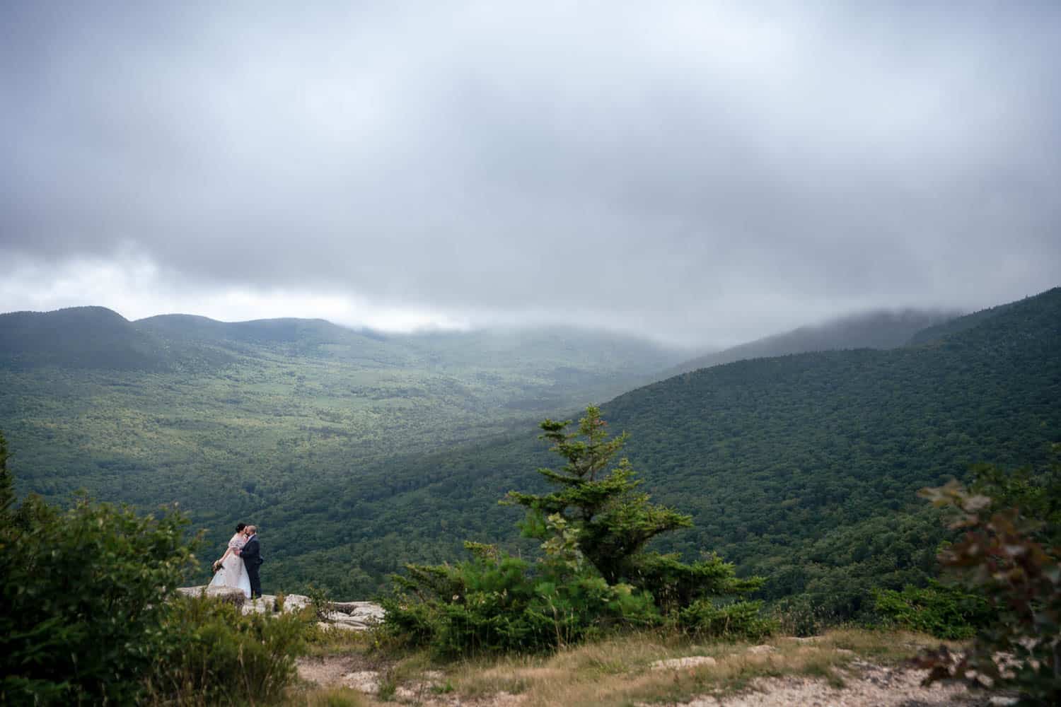 A couple in wedding attire stands on a rocky ledge embracing, overlooking a vast, green mountainous landscape under a cloudy sky.