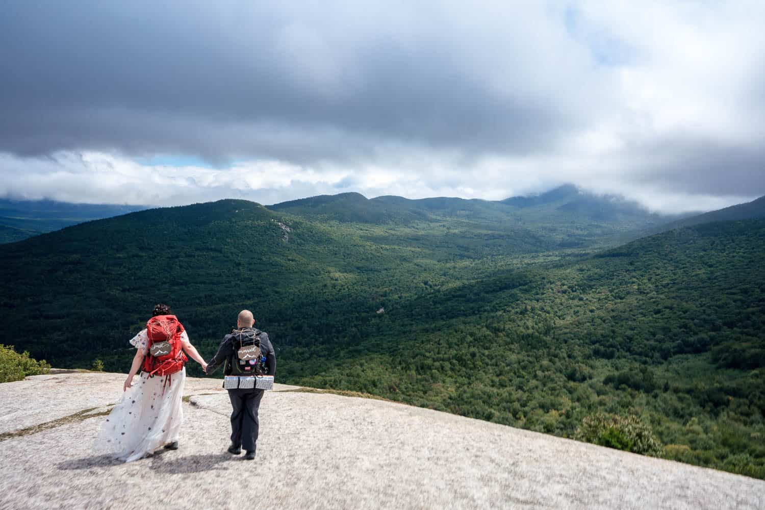 Two people wearing backpacks hold hands while standing on a rocky surface with a vast, green mountainous landscape and a cloudy sky in the background.