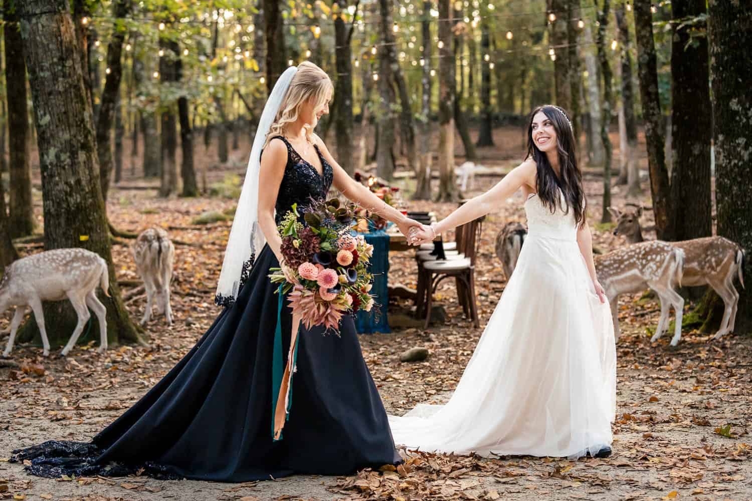 Two brides in black and white gowns hold hands in a forest, surrounded by deer, with a decorated table in the background.