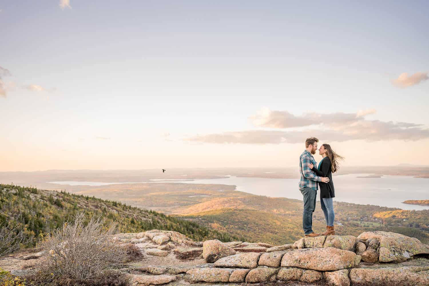 A couple stands on a rocky cliff with expansive views of a lake and hills in the background under a partly cloudy sky.