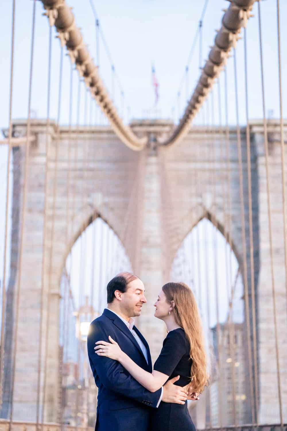 A man and a woman embrace under the cables of the Brooklyn Bridge, looking into each other's eyes. Both are dressed in formal attire.