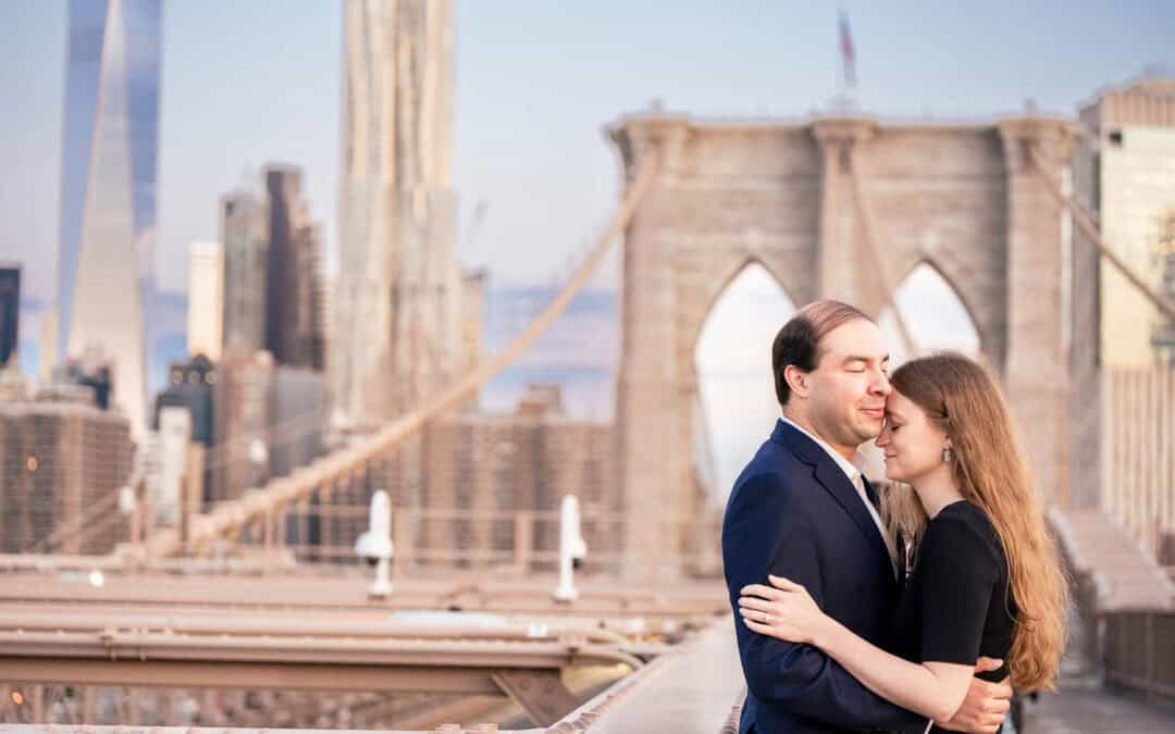 A couple embraces on a city bridge with the skyline and tall buildings visible in the background.
