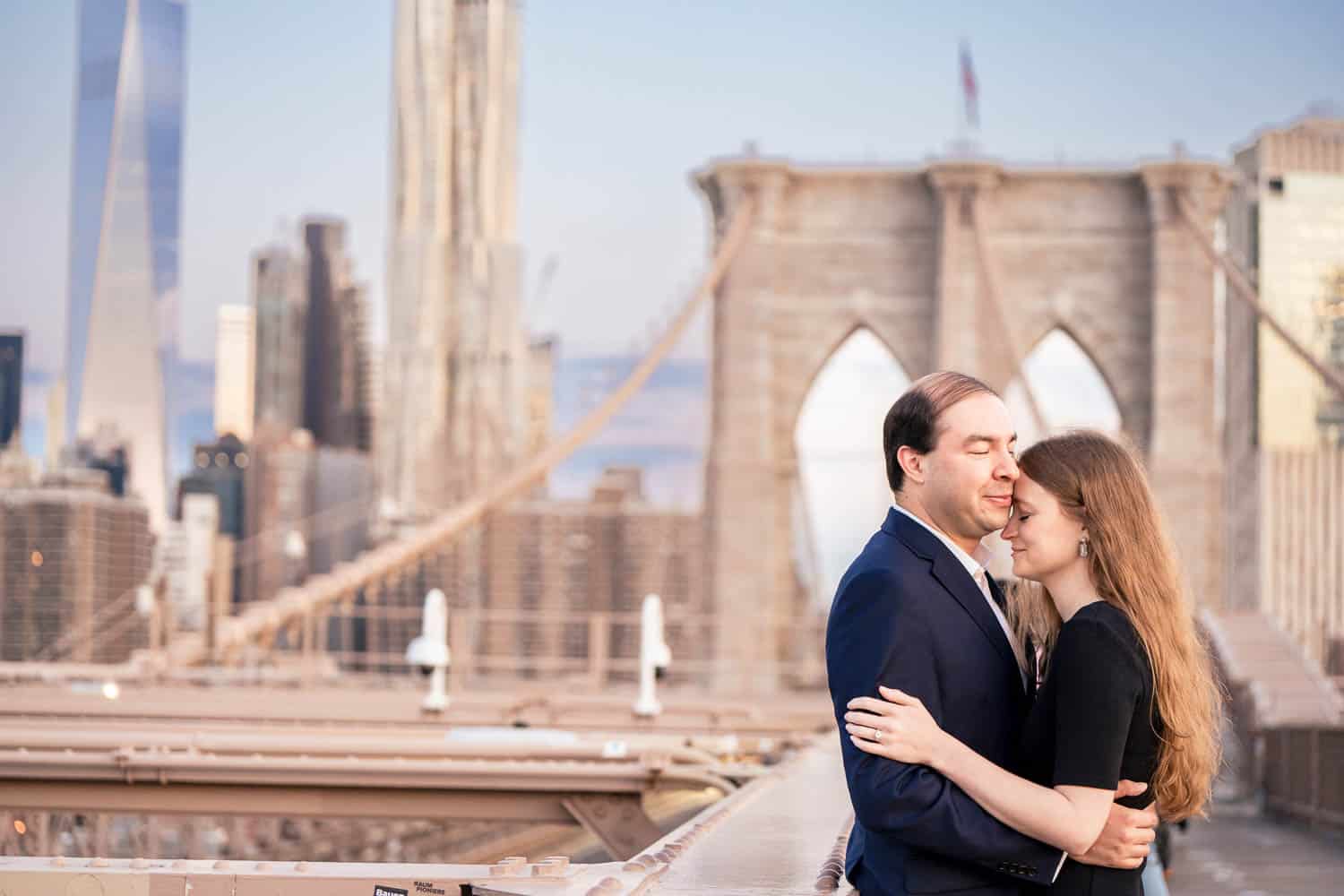 A couple embraces on a city bridge with the skyline and tall buildings visible in the background.