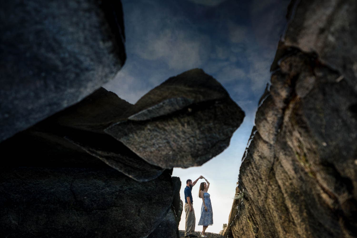 A couple's reflection is seen in a water puddle among large rocks; the man twirls the woman as if they are dancing.