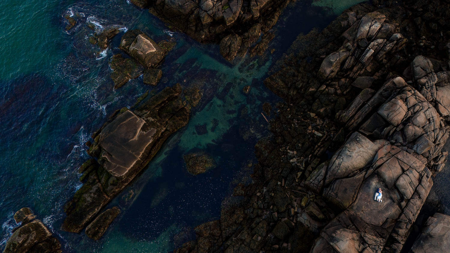 Aerial view of a person lying on large rocks near a rocky shoreline with waves gently hitting the rocks, surrounding water varying in shades of blue and green.