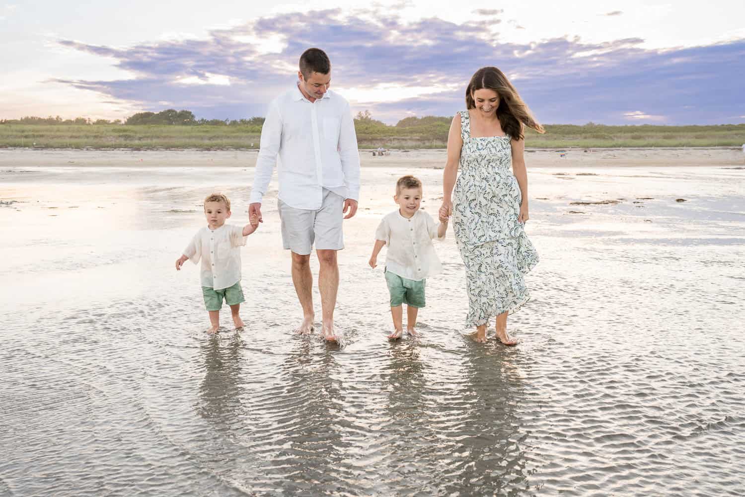 A family of 2 adults and 2 children, walk barefoot along a beach at sunset. The adults hold hands with the children, who are wearing matching outfits.