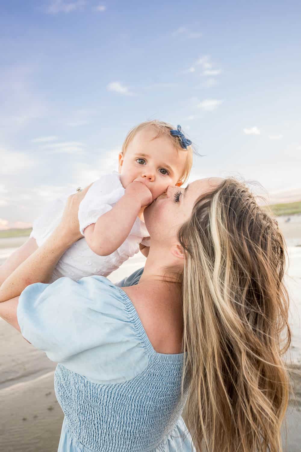 A woman with long, flowing hair kisses a baby who is sucking her thumb. They are on a beach with a clear blue sky in the background.