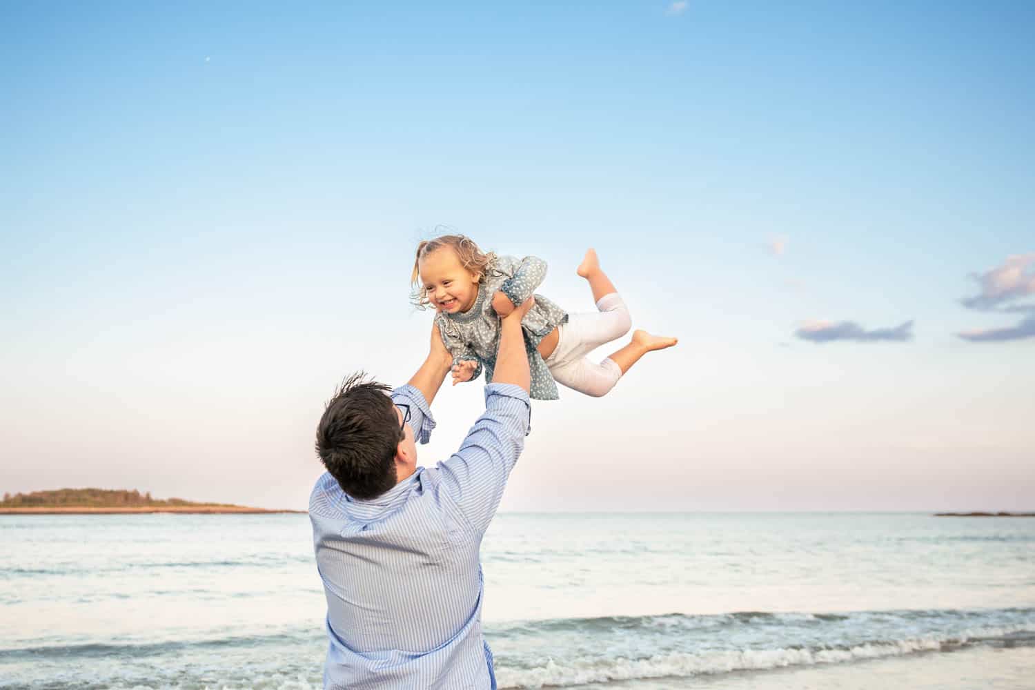 An adult lifts a smiling child into the air on a beach with calm water and a clear sky in the background.