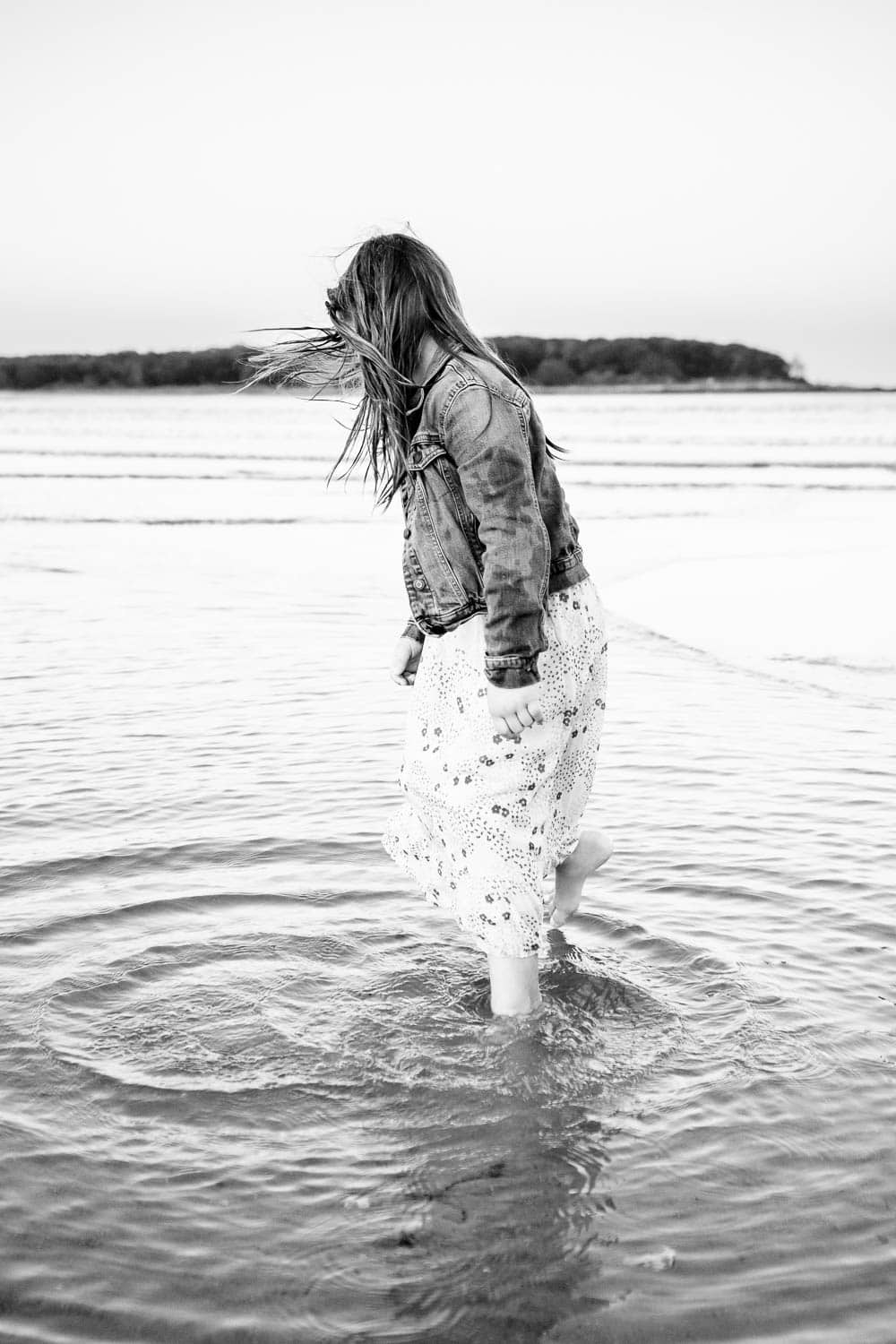 A woman wearing a denim jacket and floral skirt walks through shallow water at a beach, creating ripples around her feet.