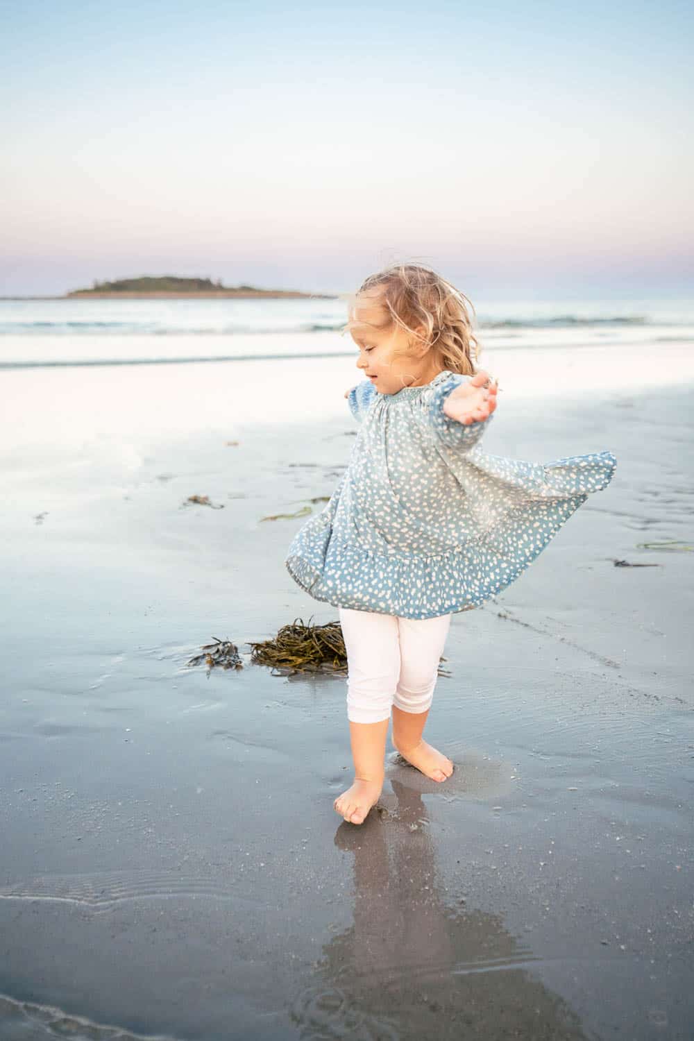 A young child joyfully twirls barefoot on a sandy beach, wearing a light blue dress with white polka dots and white leggings.