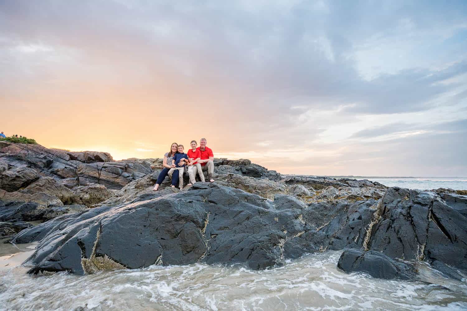 Four people sit on rocky terrain by the ocean at sunset, with waves splashing against the rocks and clouds in the sky.