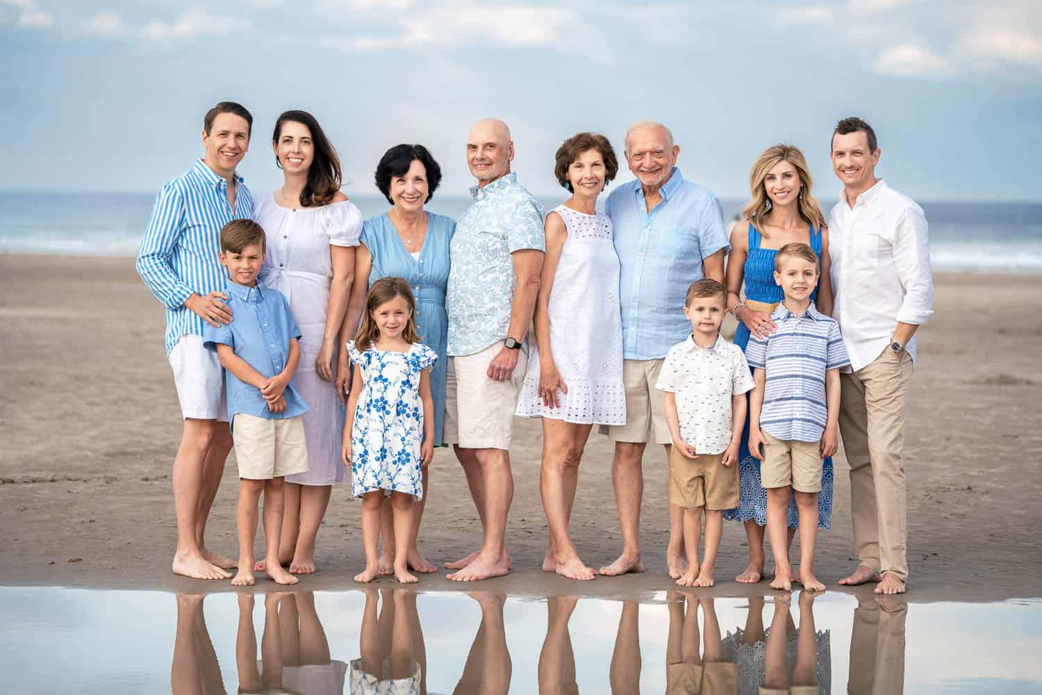 Ten casually dressed adults and three children stand in a line on a beach, with the ocean in the background and are reflected in the wet sand.