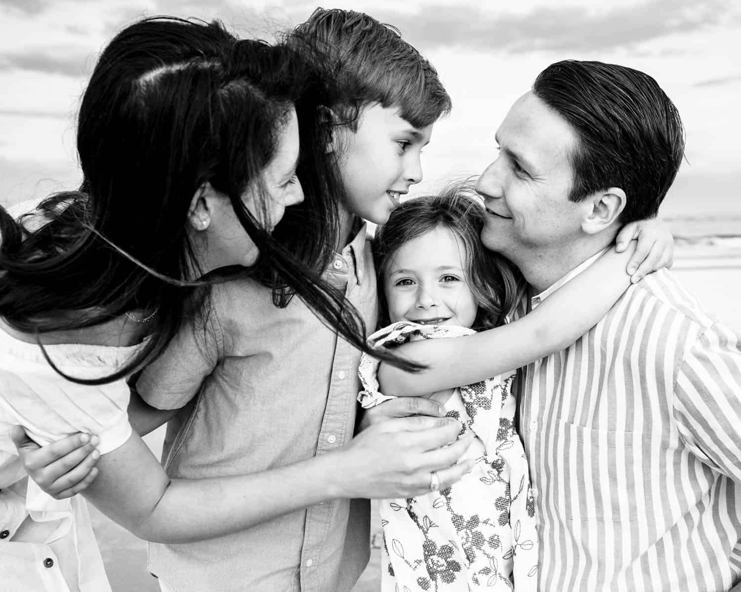 A family of four shares a close embrace, with the parents smiling at their young son and daughter on a beach. The image is in black and white.