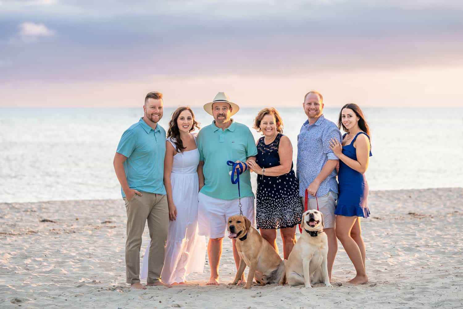 A group of six people and two dogs pose for a photo on a beach with the ocean and a pastel-colored sky in the background.