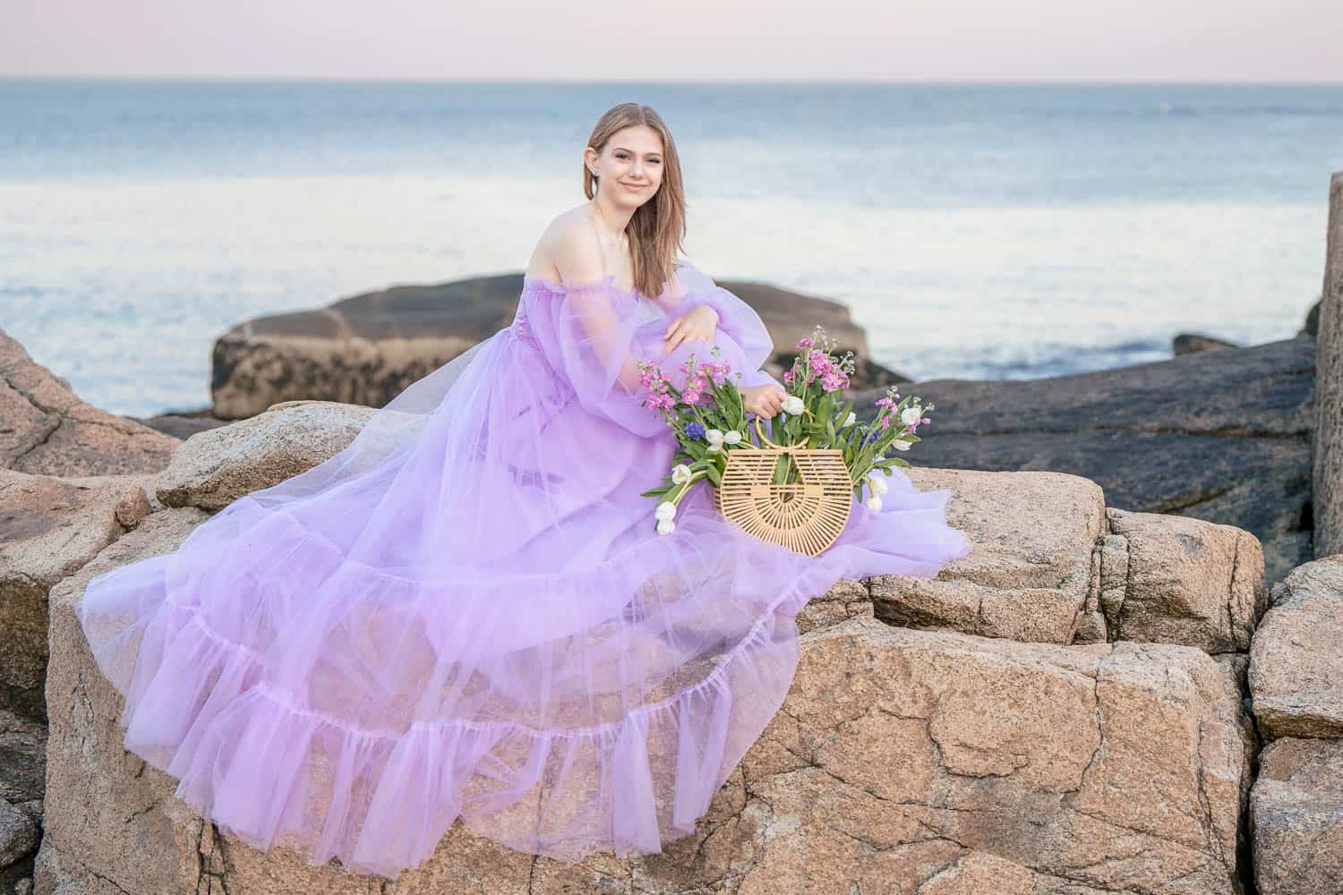 A woman in a flowing purple dress sits on large rocks by the sea, holding a basket of flowers.